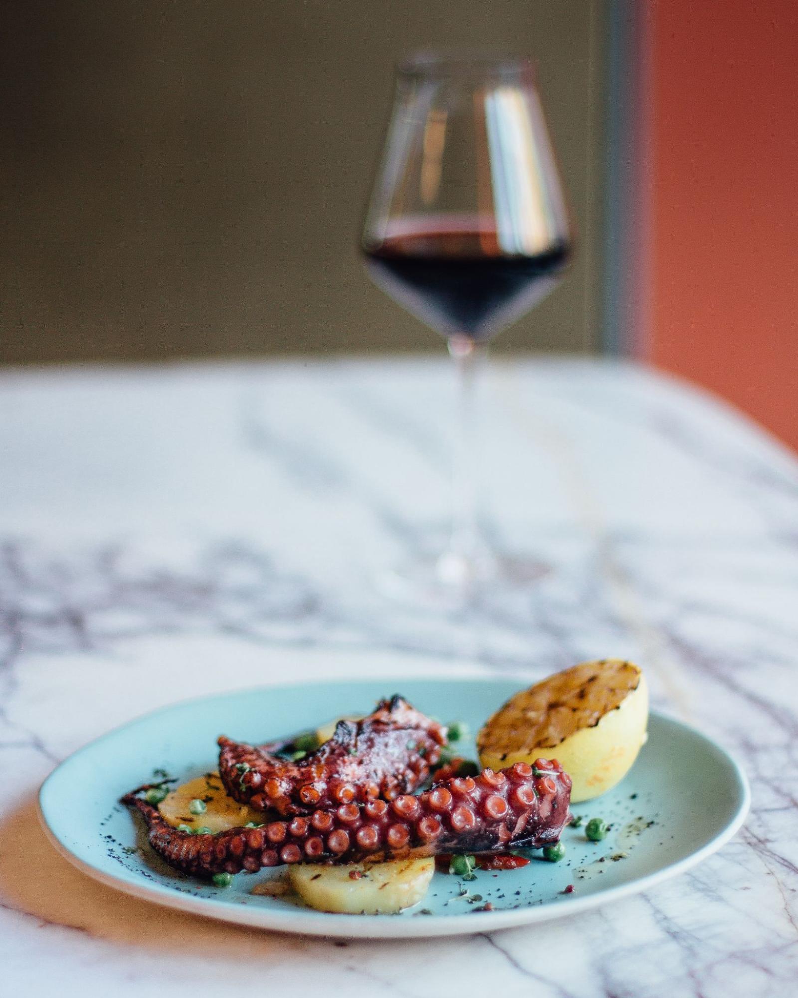 Plated octopus with sides and a grilled lemon half and a glass of red wine